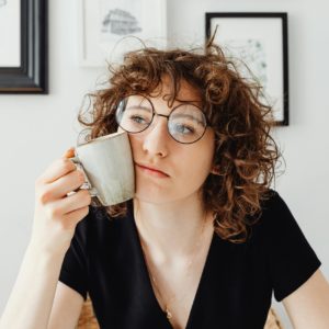 A thoughtful woman with curly hair and glasses holding a coffee mug in a modern room.